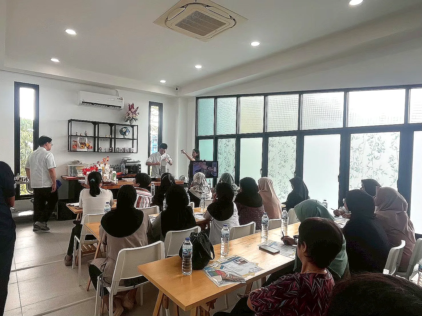 ผู้เข้าร่วมนั่งเรียนที่โต๊ะและมองไปยังพื้นที่สาธิตกับจอแสดงภาพด้านหน้าห้อง